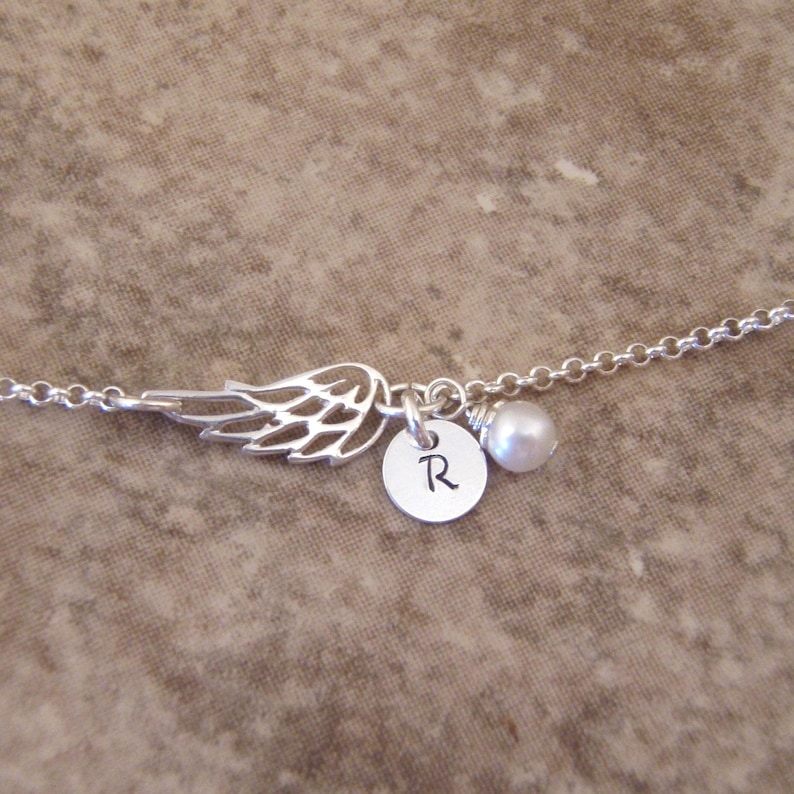 May include: A silver-toned necklace featuring a delicate wing charm, a small circular pendant with the letter "R", and a single pearl. The chain is fine and the design is simple and elegant.