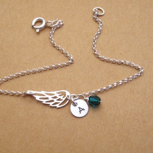 May include: A silver-colored bracelet featuring a delicate wing charm, a small round pendant with the letter "A", and a dark green crystal bead. The bracelet is made of a fine chain and has a clasp closure.