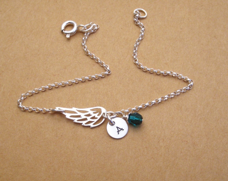 May include: A silver-colored bracelet featuring a delicate wing charm, a small round pendant with the letter "A", and a dark green crystal bead. The bracelet is made of a fine chain and has a clasp closure.