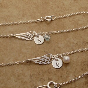May include: Silver necklaces with wing charms, personalized with initialed round pendants and small accent beads. The necklaces are displayed on a neutral brown surface. The wing charms are detailed and delicate.