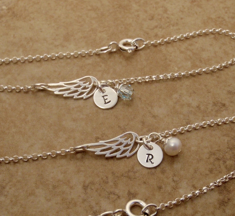 May include: Silver necklaces with wing charms, personalized with initialed round pendants and small accent beads. The necklaces are displayed on a neutral brown surface. The wing charms are detailed and delicate.