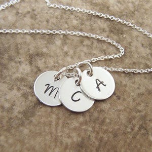 May include: A silver necklace with three round pendants, each engraved with a single letter: M, C, and A. The pendants are connected to a delicate chain, creating a personalized jewelry piece.