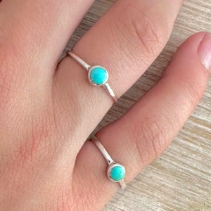 May include: Two silver rings with round, bright turquoise stones. The rings are simple bands with a single stone set in the center. The rings are on a person's fingers, showcasing the jewelry.
