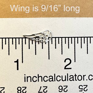 May include: A small, silver-colored wing charm, measuring 9/16 inches long, is displayed on a ruler. The wing has a detailed, open design. The text "Wing is 9/16" long" is above the wing.