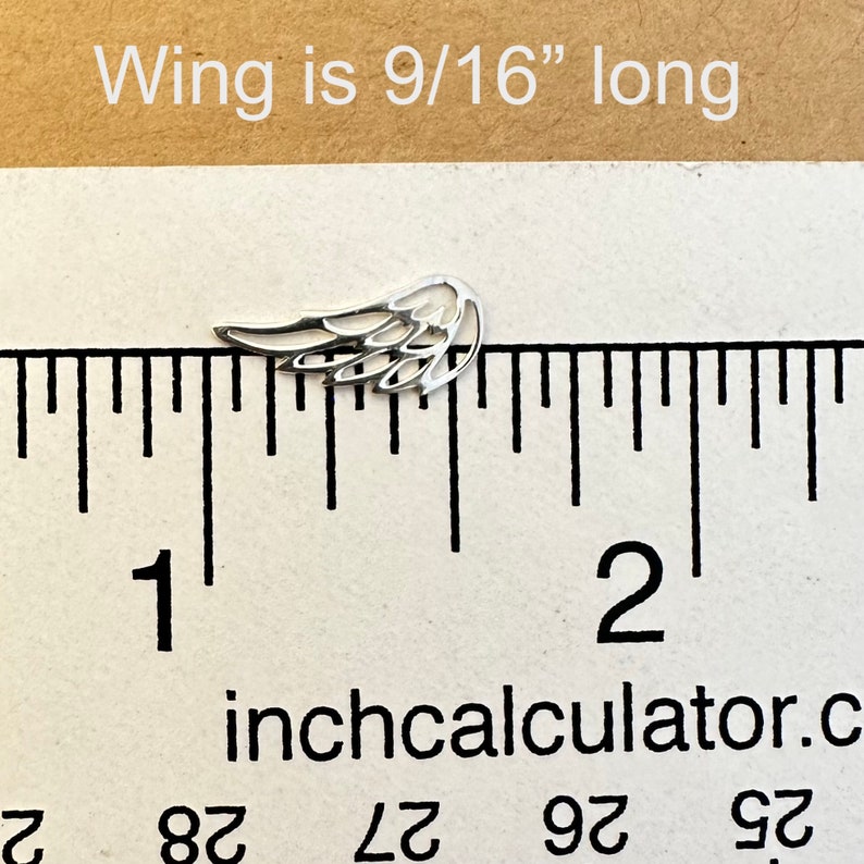 May include: A small, silver-colored wing charm, measuring 9/16 inches long, is displayed on a ruler. The wing has a detailed, open design. The text "Wing is 9/16" long" is above the wing.