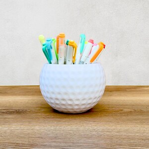 May include: A white ceramic pencil holder shaped like a golf ball, containing a selection of colourful markers.