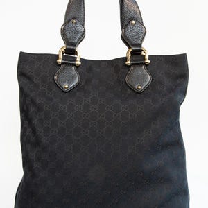 May include: Black tote bag with a repeating pattern. The bag has two handles with gold-colored hardware and leather accents. The bottom corners of the bag are reinforced with leather.