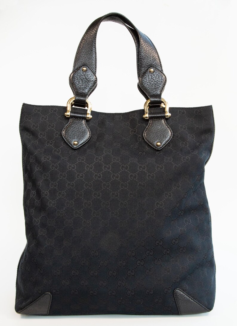 May include: Black tote bag with a repeating pattern. The bag has two handles with gold-colored hardware and leather accents. The bottom corners of the bag are reinforced with leather.