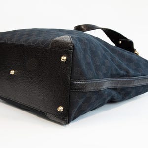 May include: Black handbag with a textured leather base and gold-colored studs. The bag's body is a dark blue fabric with a repeating pattern. A black leather handle is visible at the top.