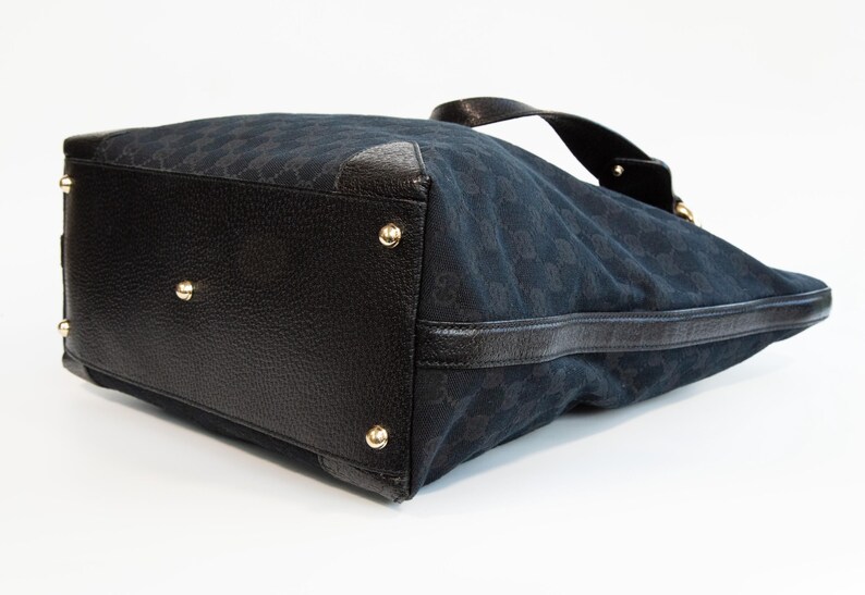 May include: Black handbag with a textured leather base and gold-colored studs. The bag's body is a dark blue fabric with a repeating pattern. A black leather handle is visible at the top.