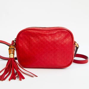May include: A bright red leather crossbody bag featuring an embossed repeating logo pattern across the surface. The bag includes a matching red leather tassel attached to a bamboo-style hardware accent and a thin adjustable shoulder strap.