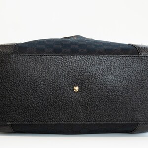 May include: Bottom view of a black handbag with a textured leather base and gold-toned metal feet. The bag features a patterned fabric top section and a central gold-toned stud.