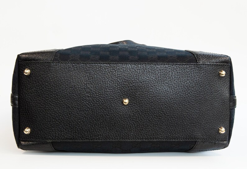 May include: Bottom view of a black handbag with a textured leather base and gold-toned metal feet. The bag features a patterned fabric top section and a central gold-toned stud.