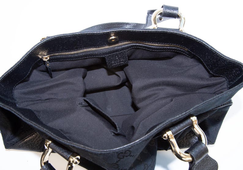 May include: Black tote bag with gold-tone hardware. The interior is black with a zippered pocket. The bag has two handles and a magnetic snap closure. The exterior features a subtle pattern.