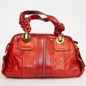 May include: A red leather handbag with plaited handles and gold-tone hardware. The bag has a structured design with vertical seams and a zip closure. The handles are attached with gold rings.