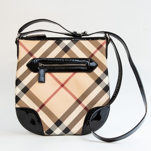 May include: A beige crossbody bag with a classic plaid pattern in black, brown, and red. The bag features a black patent leather trim, a zippered front pocket, and a long, adjustable strap.
