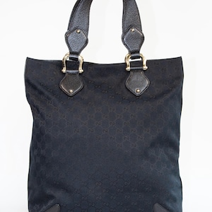 May include: Black tote bag with a repeating pattern. The bag has two black leather handles attached with gold-colored hardware. The bag's corners are reinforced with black leather patches. The bag is set against a white background.