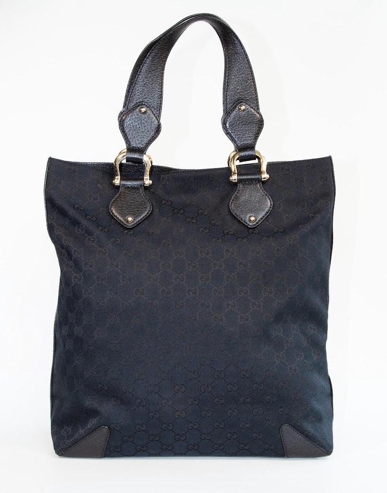 May include: Black tote bag with a repeating pattern. The bag has two black leather handles attached with gold-colored hardware. The bag's corners are reinforced with black leather patches. The bag is set against a white background.