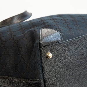 May include: Close-up of a black handbag with a repeating pattern. The bag features black leather trim and gold-colored metal studs. The fabric has a subtle sheen.