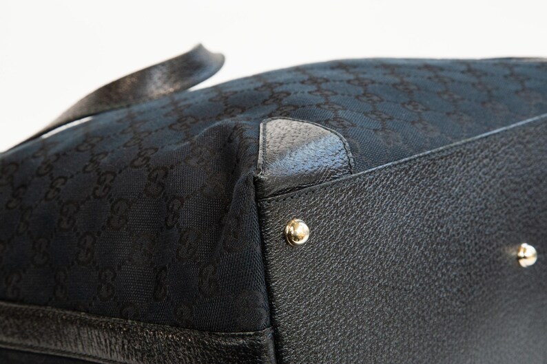 May include: Close-up of a black handbag with a repeating pattern. The bag features black leather trim and gold-colored metal studs. The fabric has a subtle sheen.