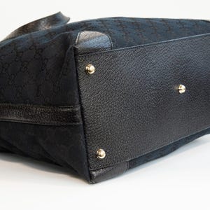 May include: Black tote bag with a repeating pattern. The bag features black leather trim and gold-colored metal studs on the bottom. The bag has a structured shape and a handle.