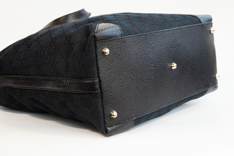 May include: Black tote bag with a repeating pattern. The bag features black leather trim and gold-colored metal studs on the bottom. The bag has a structured shape and a handle.