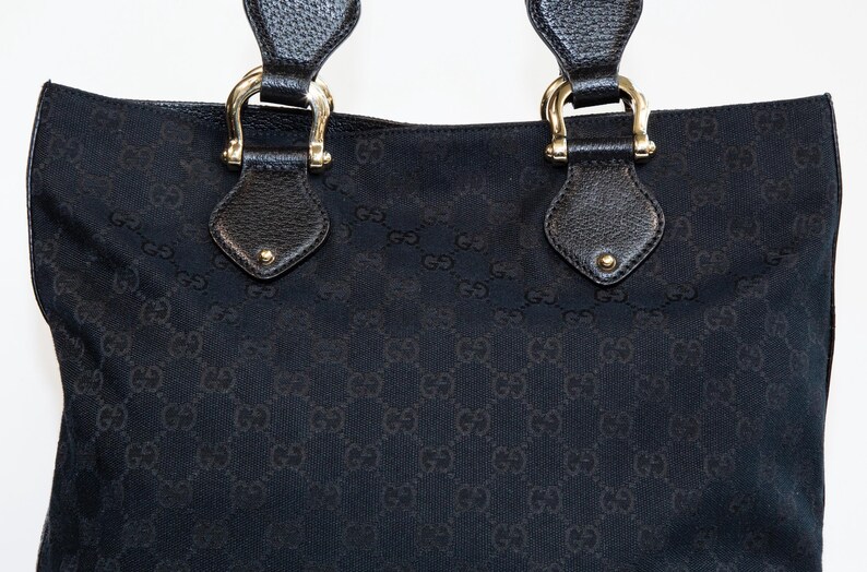 May include: Black tote bag with a repeating pattern. The bag has two handles with gold-colored hardware and leather accents. The bag's material has a subtle texture, and the overall design is classic and elegant.