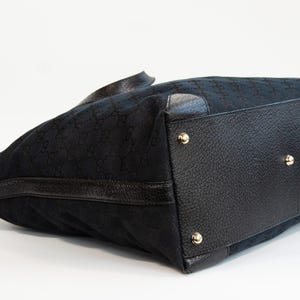 May include: Black tote bag with a repeating pattern. The bag features black leather trim and gold-colored studs on the bottom. The bag is made of a textured fabric.