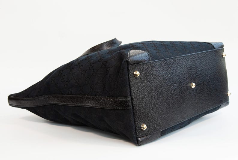 May include: Black tote bag with a repeating pattern. The bag features black leather trim and gold-colored studs on the bottom. The bag is made of a textured fabric.