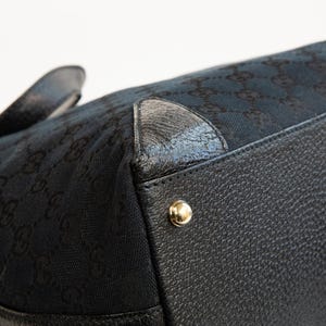 May include: Close-up of a black handbag with a textured leather base and gold-colored metal studs. The bag's upper section features a dark fabric with a repeating pattern. The handle is visible in the background.