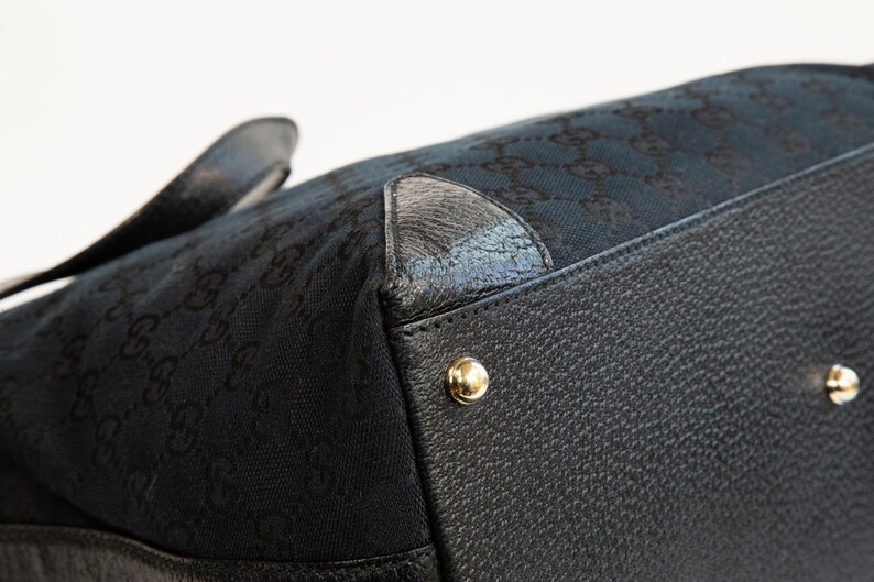 May include: Close-up of a black handbag with a textured leather base and gold-colored metal studs. The bag's upper section features a dark fabric with a repeating pattern. The handle is visible in the background.