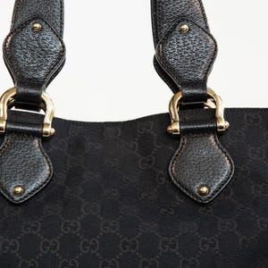 May include: Close-up of a black tote bag with a repeating pattern. The bag features black leather handles with gold-colored hardware. The handles are attached to the bag with gold-colored metal rings.