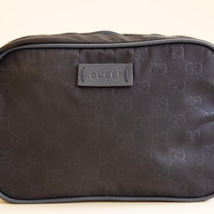 Può includere: Una borsa da viaggio rettangolare blu navy con un motivo in rilievo ripetuto. La borsa ha un manico e finiture in pelle e una piccola etichetta in pelle con il nome del marchio "Gucci".