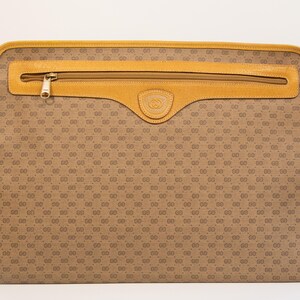 May include: A brown and tan Gucci briefcase with a repeating GG logo pattern. The briefcase has a tan leather trim, a zippered pocket, and a gold-tone zipper pull. The Gucci logo is embossed on a leather patch.