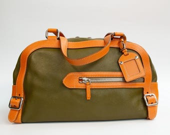May include: An olive green leather handbag featuring bright orange leather trim, two top handles, a horizontal zippered front pocket with a silver-tone metal zipper, and two side buckles. A matching orange leather luggage tag is attached.