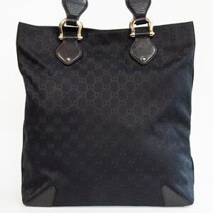 May include: Black tote bag with a repeating pattern. The bag has two handles with gold-colored hardware and leather accents. The bottom corners are reinforced with dark leather. The bag is made of a woven material.