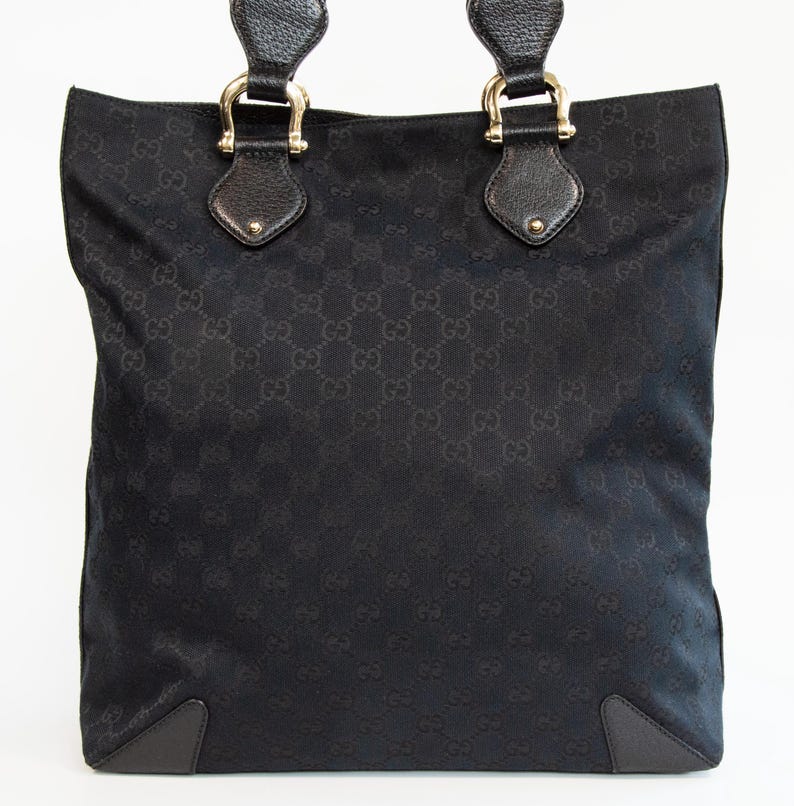 May include: Black tote bag with a repeating pattern. The bag has two handles with gold-colored hardware and leather accents. The bottom corners are reinforced with dark leather. The bag is made of a woven material.