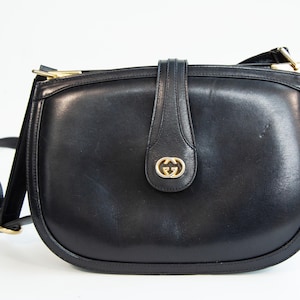 Puede incluir: Un bolso de hombro de cuero negro con silueta redondeada, que presenta un cierre de correa vertical central y un emblema con el logotipo GG en metal dorado. El bolso incluye una correa de hombro de cuero negro a juego con herrajes dorados.