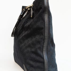 May include: Black tote bag with a repeating pattern. The bag features a leather trim and gold-colored hardware. The bag has a shoulder strap and a structured shape. The bag is made of a woven material.