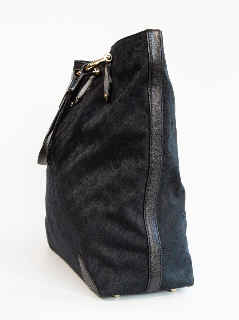 May include: Black tote bag with a repeating pattern. The bag features a leather trim and gold-colored hardware. The bag has a shoulder strap and a structured shape. The bag is made of a woven material.