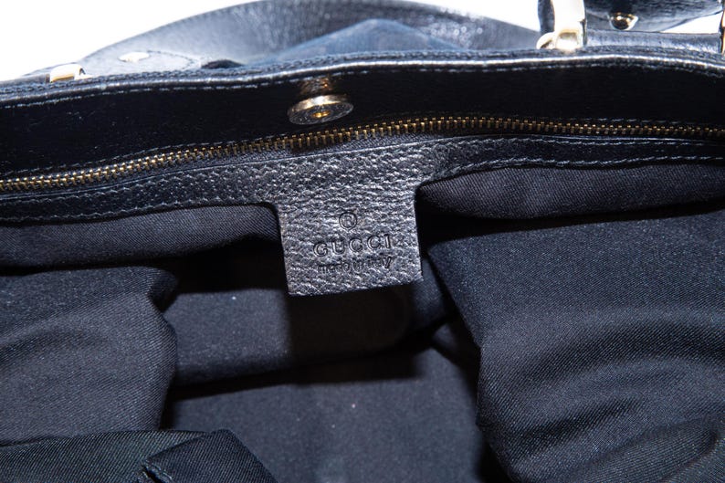 May include: Close-up of a black leather handbag with a gold zipper and hardware. The interior is lined with black fabric. A leather tag with the "GUCCI" logo is visible.