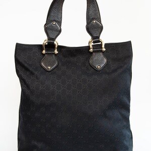 May include: Black tote bag with leather handles and gold-tone hardware. The bag features a repeating pattern across the fabric. The handles are attached with gold-tone metal rings. The bag has a rectangular shape.