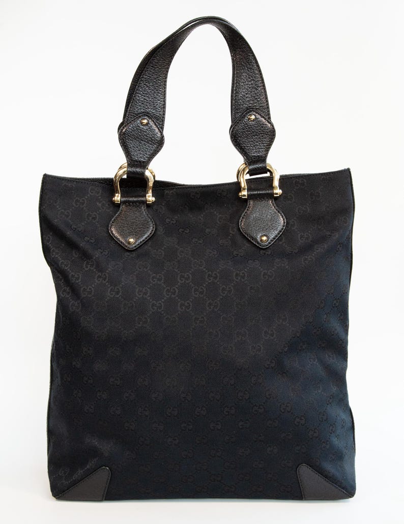 May include: Black tote bag with leather handles and gold-tone hardware. The bag features a repeating pattern across the fabric. The handles are attached with gold-tone metal rings. The bag has a rectangular shape.