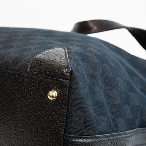 May include: Close-up of a black handbag with a repeating pattern. The bag features black leather accents, gold-colored hardware, and a black handle. The bag's material has a subtle sheen.