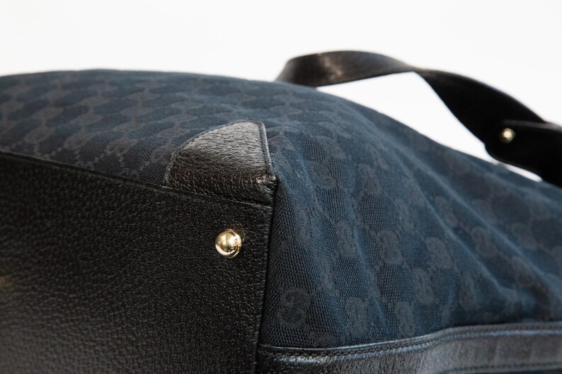 May include: Close-up of a black handbag with a repeating pattern. The bag features black leather accents, gold-colored hardware, and a black handle. The bag's material has a subtle sheen.