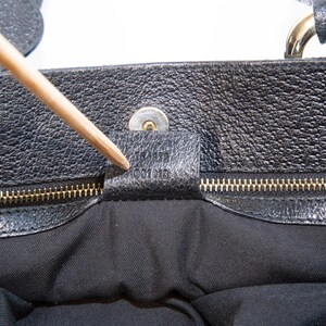 May include: Close-up of a black leather handbag. The bag features a textured surface, a gold-colored zipper, and a small leather tag with embossed text. A wooden pointer is touching the tag.