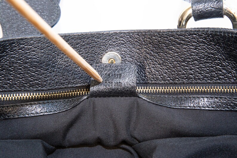 May include: Close-up of a black leather handbag. The bag features a textured surface, a gold-colored zipper, and a small leather tag with embossed text. A wooden pointer is touching the tag.