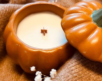Könnte beinhalten: Ein kürbisförmiger Kerzenbehälter aus Keramik mit glänzend orangefarbener Oberfläche und abnehmbarem Deckel. Die Kerze enthält weißes Wachs und einen kreuzförmigen Holzdocht. Der Deckel ist mit einem grünen Stiel versehen.