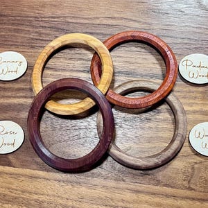 May include: Four wooden bangles in shades of yellow, red-brown, dark purple-brown, and light brown are displayed. Small wooden tags with the words "Canary Wood", "Padauk Wood", "Rose Wood", and "Walnut Wood" are also present. The bangles are on a wooden surface.