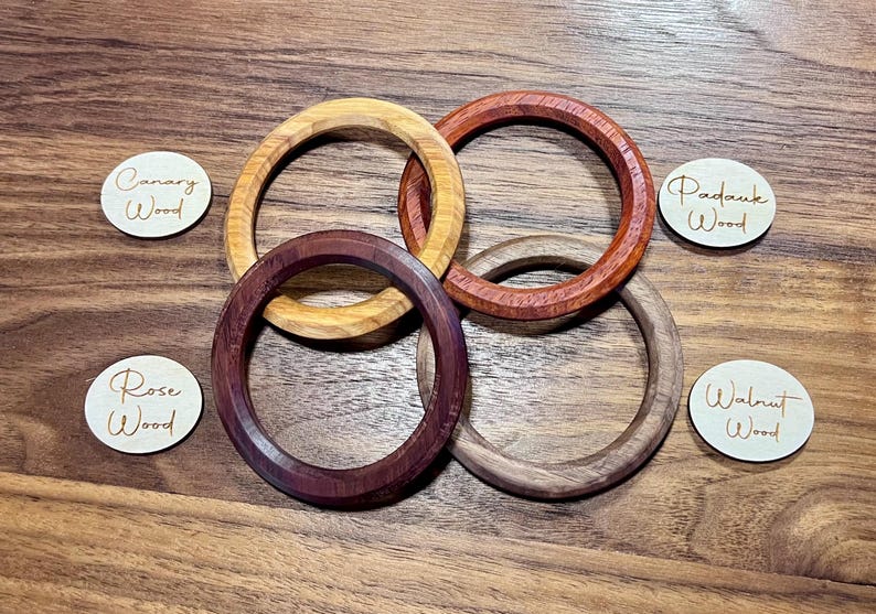 May include: Four wooden bangles in shades of yellow, red-brown, dark purple-brown, and light brown are displayed. Small wooden tags with the words "Canary Wood", "Padauk Wood", "Rose Wood", and "Walnut Wood" are also present. The bangles are on a wooden surface.